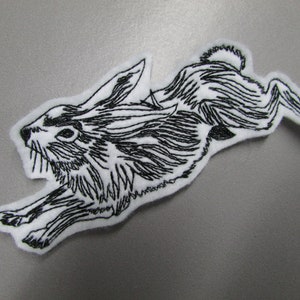 May include: Black and white embroidered patch of a rabbit running. The rabbit is detailed with fur and has a long tail.