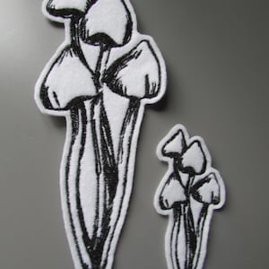 Puede incluir: Dos parches de tela blanca con una ilustración en tinta negra de un grupo de setas. El parche más grande muestra tres setas con tallos y sombreros detallados, mientras que el parche más pequeño muestra un diseño similar a menor escala.