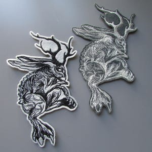 May include: Two embroidered patches featuring a jackalope with long ears and antlers. One patch has black thread on a white background, while the other features silver metallic thread on a dark gray background, both showing detailed fur textures.