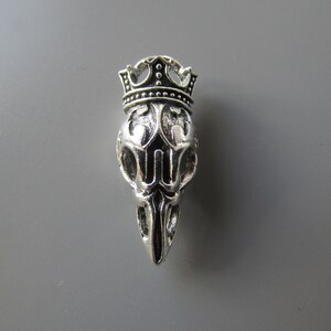 May include: Silver skull charm with a crown on top. The skull is detailed with intricate carvings and the crown has black accents.