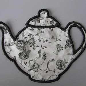 May include: A black and white teapot-shaped tea cosy with a floral pattern. The cosy is made of fabric and has a black trim.