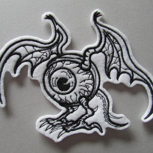 May include: A white embroidered patch featuring a stylized eye with wings and clawed feet. The design is outlined in black, with detailed wing patterns and eye features. The patch has a fantasy or gothic theme.