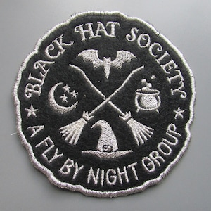 Może przedstawiać: Naszywka haftowana w kolorze czarnym i białym z tekstem "Black Hat Society A Fly By Night Group" i obrazami nietoperza, kotła, rosnącego księżyca i dwóch skrzyżowanych miotł.