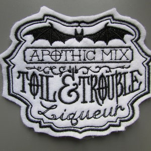 Puede incluir: Parche blanco bordado con puntadas negras. El parche presenta un murciélago, una vid decorativa y el texto "Apothecary Mix", "Toil & Trouble" y "Liqueur".