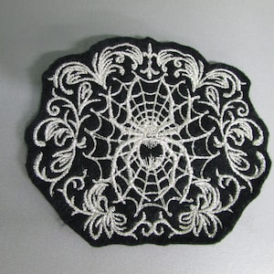 May include: A black felt patch with a silver embroidered spider web and spider in the center. The spider web is surrounded by a silver embroidered floral design.