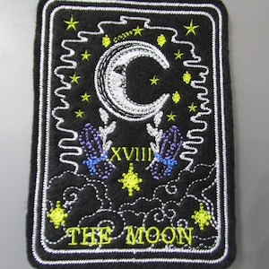 May include: Black felt patch with white embroidery of the The Moon tarot card. The card features a crescent moon, two towers, a dog, and a crab. The card number XVIII is in Roman numerals.
