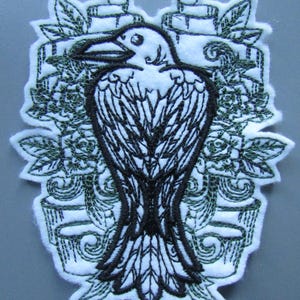 May include: Black and green embroidered patch featuring a raven with a detailed design. The raven is surrounded by floral and scrollwork designs.