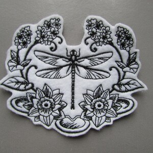 Puede incluir: Parche bordado en blanco y negro con una libélula con las alas extendidas, rodeada de flores y hojas estilizadas.