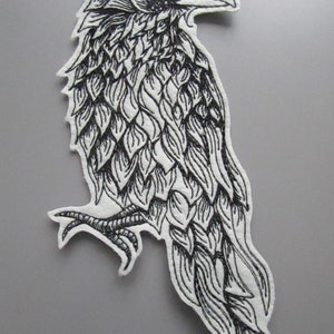 May include: Black and white embroidered patch of a crow with detailed feathers. The crow is facing right and has a long beak. The patch is made of felt and has a white background.