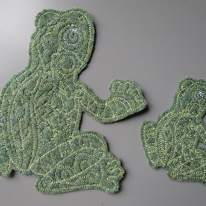 May include: Two green frog-shaped fabric patches featuring intricate, textured embroidery in shades of olive and lime green. The larger patch is positioned on the left, while a smaller, matching frog patch is located to the right of it.