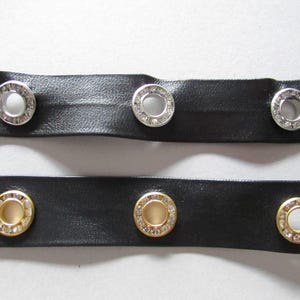 May include: Close-up of two black leather strips with decorative eyelets. The top strip has silver eyelets with clear rhinestones, while the bottom strip features gold eyelets with clear rhinestones. The eyelets are evenly spaced.