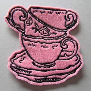 Könnte beinhalten: Rosa Filz-Applikation mit einem Design aus einer gestapelten Teetasse und Untertasse mit schwarzen Stickkonturen. Die Teetasse hat florale Details und einen gebogenen Henkel. Die Untertasse ist rund mit einem gewellten Rand. Für Bastel- und Nähprojekte.