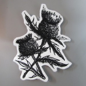 May include: A black and white embroidered patch featuring a thistle design. The patch has a white background and a detailed black outline of the thistle flowers and leaves. The patch is an irregular shape.