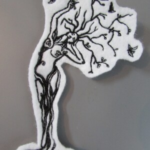 May include: Black and white embroidered patch of a woman's figure with branches growing from her head and body, resembling a tree. The patch is on a white background.
