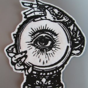May include: Black and white embroidered patch depicting a hand holding a crystal ball with an eye inside. The eye has a black pupil and white iris. The hand is adorned with intricate details.