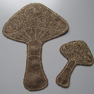 May include: Two mushroom-shaped fabric appliqués featuring a brown and black herringbone pattern. Each piece is outlined with gold metallic stitching that also forms decorative swirl patterns on the mushroom caps and vertical lines on the stems.