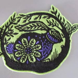 Könnte beinhalten: Gestickter Aufnäher mit einem Kaninchen-Design. Der Aufnäher hat eine schwarze Kaninchensilhouette mit lila Akzenten, umgeben von einem grünen, blattförmigen Rand. Das Design beinhaltet eine Blume und eignet sich für Kleidung oder Accessoires.