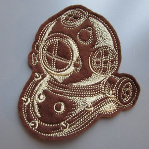Può includere: Un cerotto in feltro marrone con ricamo dorato che rappresenta un casco da sub vintage. Il casco ha un grande oblò rotondo e un oblò rotondo più piccolo sul lato. Il casco è circondato da un motivo decorativo di linee e punti a spirale.