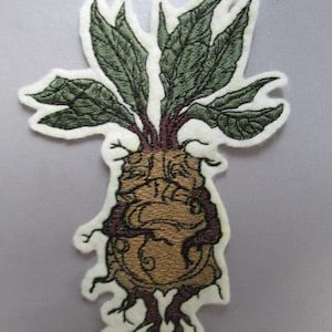 Peut inclure: Patch brodé d'une créature mythique brune et verte avec des feuilles et des racines. La créature a un corps rond et volumineux avec un visage et des bras. La créature est entourée de feuilles et de racines.
