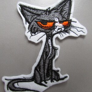 May include: Embroidered iron-on patch of a cartoon cat with a grumpy expression. The cat is black and white with orange eyes and a white patch on its nose.