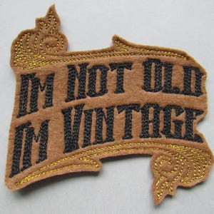 Może przedstawiać: Brązowa łatka z filcu z haftowanymi złotymi krawędziami i tekstem "I'm not old I'm vintage" w czarnym hafcie.