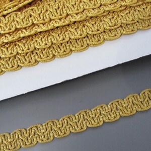 May include: Close-up of gold-colored decorative trim. The trim has a woven design with a scalloped edge. The trim is made of a shiny, metallic material and is likely used for embellishing clothing or crafts.