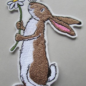 May include: Embroidered patch of a brown and white rabbit holding a daisy. The rabbit has long ears with pink inner ears and is standing upright. The patch has a white border, suitable for clothing or accessories.