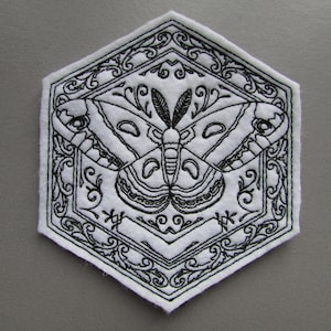 May include: A white hexagonal fabric patch with black embroidery of a moth with intricate details and a floral design around it.