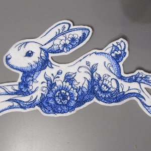 Pode incluir: Um patch bordado branco e azul em forma de coelho a correr. O coelho é decorado com padrões florais azuis. O patch é provavelmente para roupas ou acessórios.