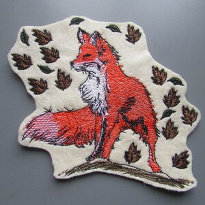 May include: Embroidered patch of a red fox with white fur on its chest and neck, standing on a brown ground with green and brown leaves falling around it.