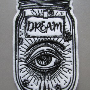May include: Black and white embroidered patch of a mason jar with the word "DREAM" on the label and an eye inside. The eye is surrounded by a sunburst design.