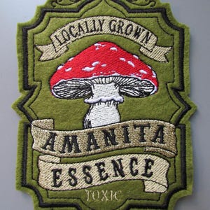 May include: An embroidered patch with an olive green felt background featuring a red and white mushroom illustration. The text reads LOCALLY GROWN, AMANITA ESSENCE, and TOXIC. The patch has a decorative scalloped border outlined in black thread.