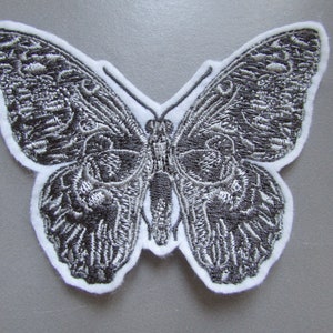May include: A silver embroidered butterfly patch with a white background. The butterfly has intricate details and a detailed wing pattern.