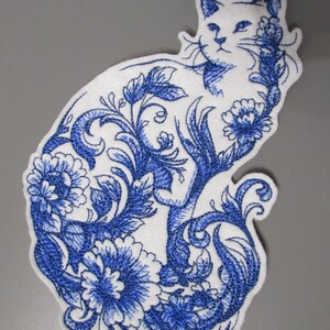 Può includere: Toppa bianca a forma di gatto con ricamo floreale blu. Il design presenta un contorno dettagliato di un gatto riempito di fiori e foglie blu vorticose, creando un pezzo decorativo e artistico.