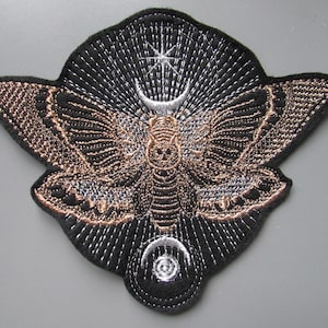 May include: Embroidered patch with a moth design, featuring a skull on its body. The patch has a black background with copper and silver thread details. The moth is surrounded by a crescent moon, star, and sunburst design.
