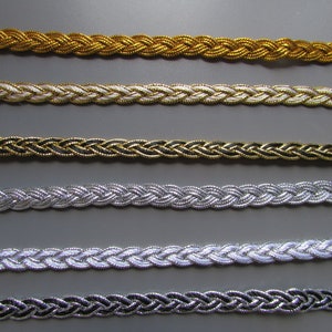 Könnte beinhalten: Sechs verschiedene geflochtene Borten in Gold, Weiß, Schwarz, Silber und Grau. Die Borten haben alle die gleiche Breite und ein ähnliches geflochtenes Muster.