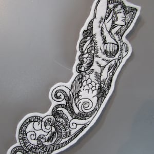 May include: Black and white embroidered patch of a mermaid with intricate details and a swirling tail.