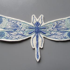 May include: Embroidered dragonfly patch in shades of blue and white. The dragonfly features intricate detailing on its wings and body. The patch is made of fabric and has a white border. The wings are a light blue with darker blue accents.
