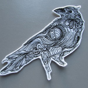 Könnte beinhalten: Schwarz-weiß gesticktes Rabendesign auf weißem Stoffhintergrund. Der Rabe ist mit komplizierten Details und wirbelnden Mustern dargestellt.