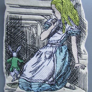 Könnte beinhalten: Gestickte Abbildung von Alice aus Alice im Wunderland, die ein blaues und weißes Kleid trägt, neben ihr ein grünes und weißes Kaninchen. Das Bild ist auf einem weißen Stoffhintergrund.