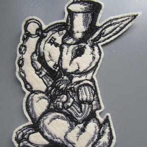May include: A black and white embroidered patch featuring a rabbit wearing a top hat and holding a pocket watch on a chain. The rabbit is depicted in a seated position with long ears, detailed fur texture, and a formal jacket, rendered in high contrast.