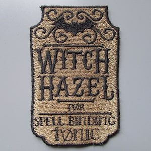 Könnte beinhalten: Ein brauner und schwarzer gestickter Aufnäher mit dem Text "Witch Hazel" und "Spell Binding Tonic". Der Aufnäher zeigt eine schwarze Fledermaus und dekorative Schnörkel.