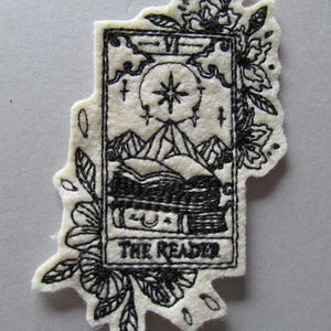 Puede incluir: Un parche bordado de color crema con un diseño de carta del tarot. La imagen en blanco y negro representa una escena de montaña, una estrella y las palabras "THE READER". El parche está rodeado de bordados florales.
