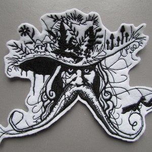 May include: An embroidered patch in white and black, depicting a stylized face with a long beard and a hat decorated with foliage. The design has intricate details and a whimsical, folk art style.