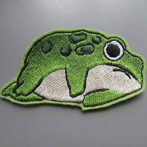 May include: Green felt frog applique with a white belly and a black eye. The frog is lying on its side with its legs tucked under its body.