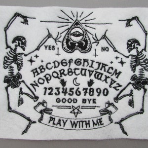 May include: Black and white embroidered patch featuring a Ouija board with two skeletons on either side. The board includes the alphabet, numbers 1-9 and 0, and the words "YES", "NO", "GOOD BYE", and "PLAY WITH ME".