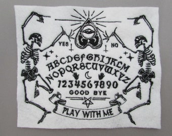 May include: Black and white embroidered patch featuring a Ouija board with two skeletons on either side. The board includes the alphabet, numbers 1-9 and 0, and the words "YES", "NO", "GOOD BYE", and "PLAY WITH ME".