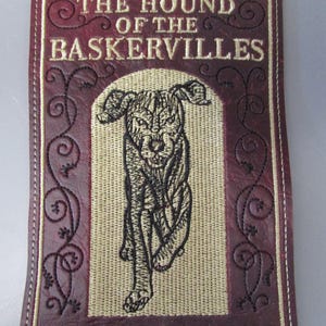 May include: Embroidered patch featuring the title "THE HOUND OF THE BASKERVILLES" and the author's name, Sir Arthur Conan Doyle. The patch has a burgundy border with a running hound illustration and decorative swirls.