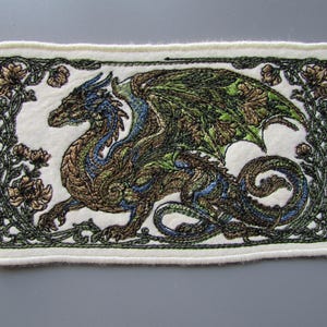 May include: A rectangular embroidered patch on a white felt background featuring a detailed dragon in shades of bronze, green, and blue. The dragon is surrounded by an intricate border of dark green embroidered vines and floral motifs.