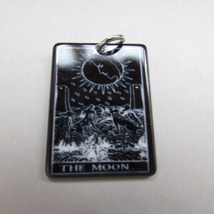 May include: Black and white tarot card pendant with a silver bail. The card depicts the Moon card from the tarot deck. The card features a crescent moon, two towers, a dog, a crab, and a path. The text "THE MOON" is printed at the bottom of the card.